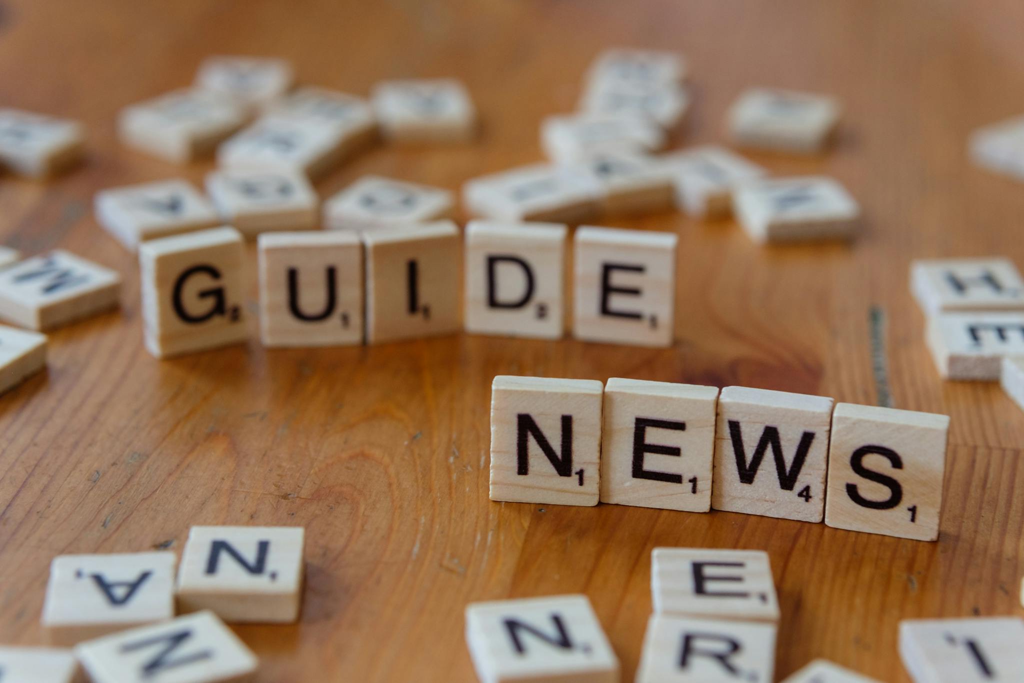 Close-up of Scrabble tiles spelling 'Guide' and 'News' on a wooden surface.